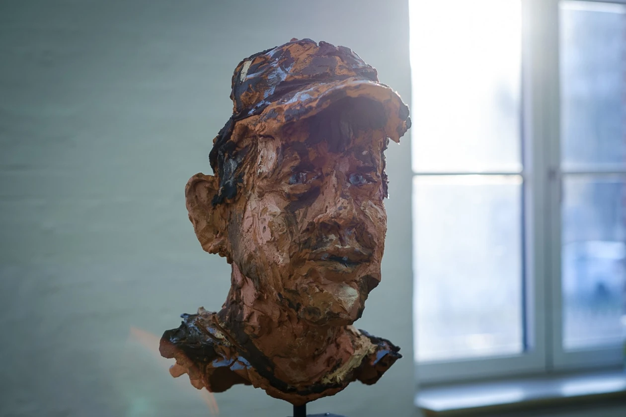 Kopfskulptur aus Ton, die ein menschliches Gesicht mit ausdrucksstarken Z&uuml;gen und unregelm&auml;&szlig;iger Oberfl&auml;che darstellt. Die Skulptur steht vor einem unscharfen Hintergrund mit einem Fenster.