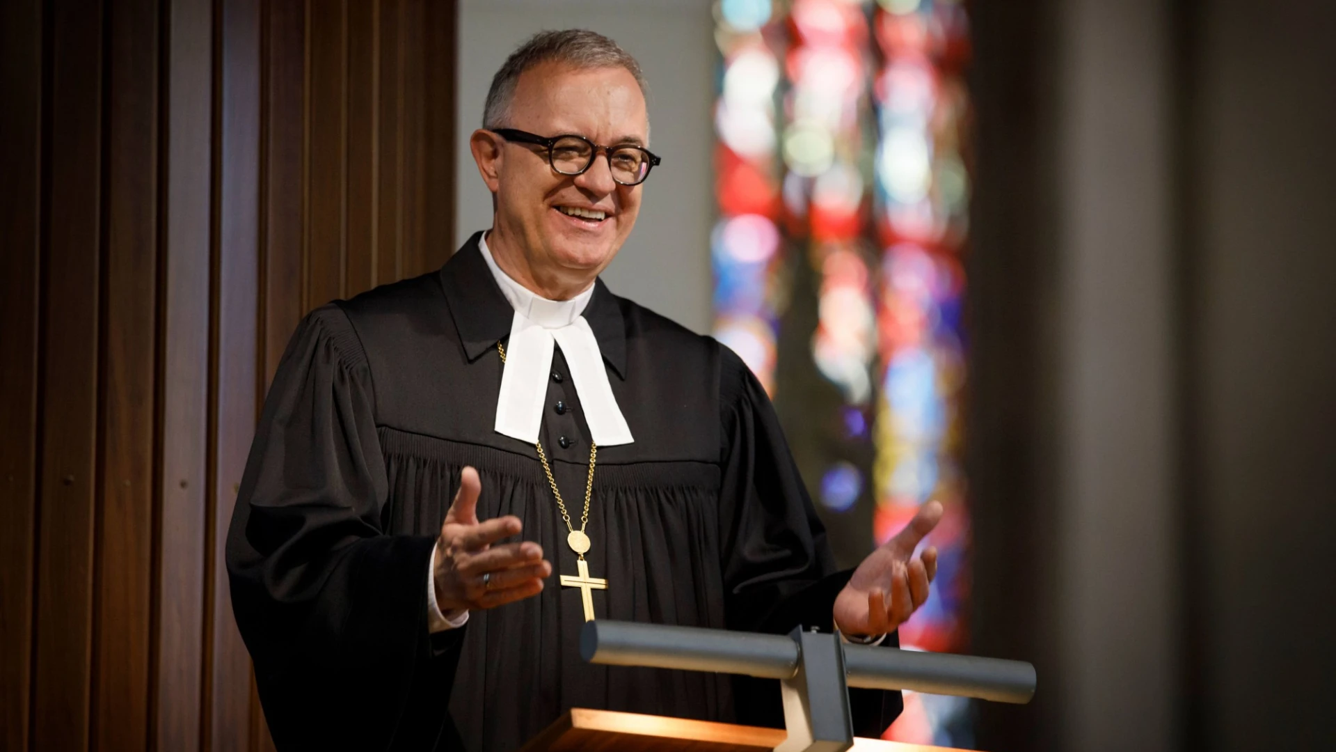 L&auml;chelnder Geistlicher in schwarzer Robe steht an einem Rednerpult. Er tr&auml;gt eine Brille und eine Kette mit einem Kreuz. Im Hintergrund sind bunte Kirchenfenster zu sehen. Eine Geste macht er mit seinen H&auml;nden.