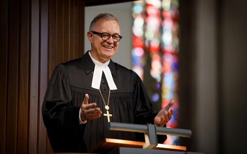 L&auml;chelnder Geistlicher in schwarzer Robe steht an einem Rednerpult. Er tr&auml;gt eine Brille und eine Kette mit einem Kreuz. Im Hintergrund sind bunte Kirchenfenster zu sehen. Eine Geste macht er mit seinen H&auml;nden.