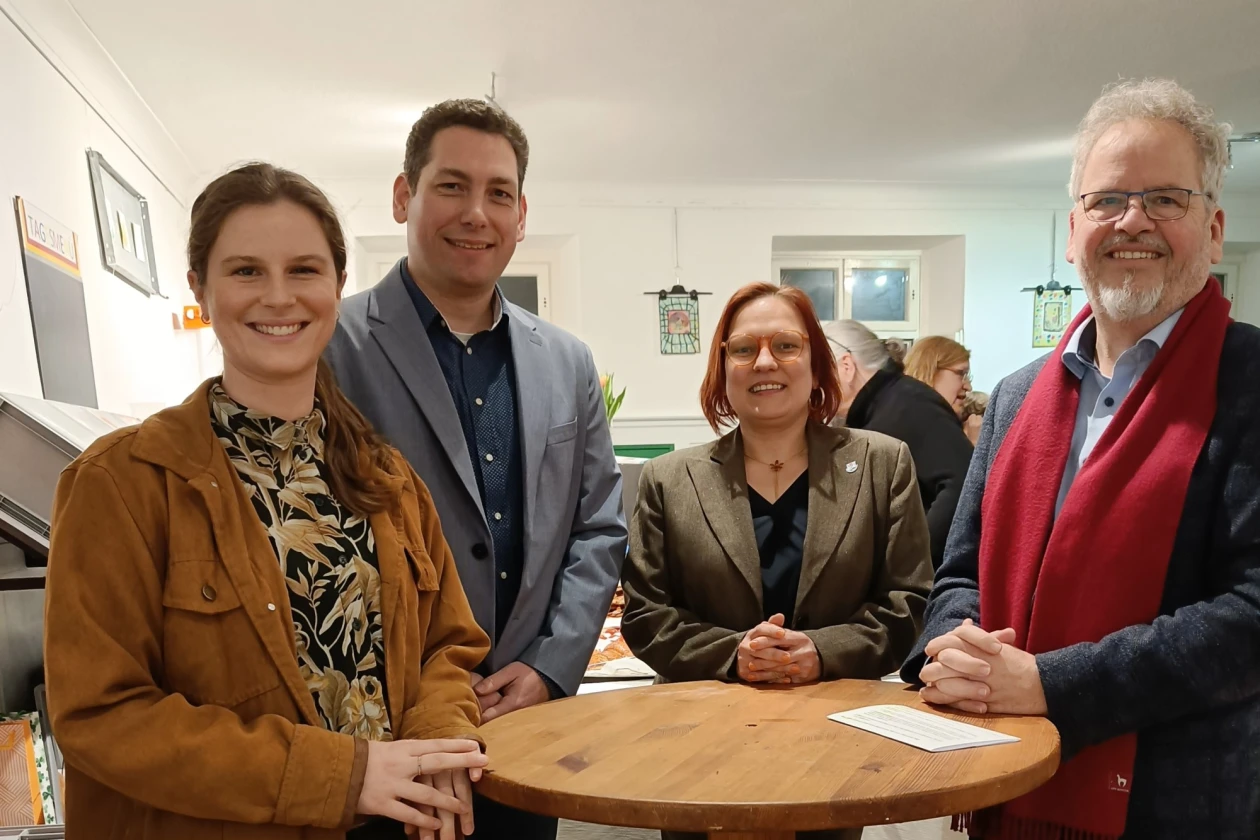 Vier Personen stehen an einem Holzstehtisch und l&auml;cheln in die Kamera. Im Hintergrund sind weitere Personen zu sehen.