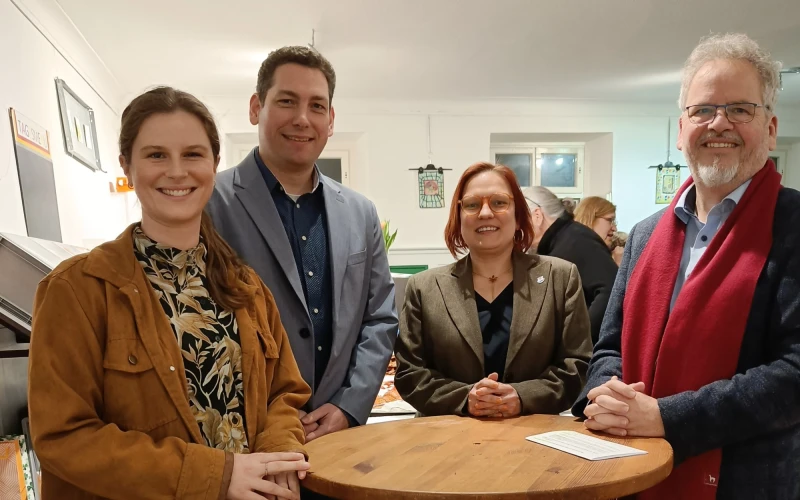 Vier Personen stehen an einem Holzstehtisch und l&auml;cheln in die Kamera. Im Hintergrund sind weitere Personen zu sehen.