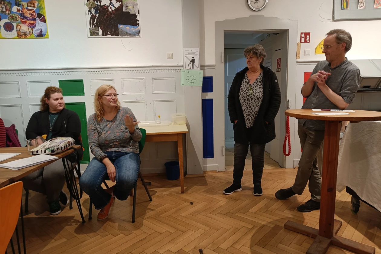 Vier Personen sind zu sehen, zwei sitzen auf St&uuml;hlen, w&auml;hrend zwei weitere stehen, einer davon an einem Stehtisch. Der Raum hat wei&szlig;e W&auml;nde, an denen Bilder h&auml;ngen. Im Hintergrund sind weitere Tische zu sehen.