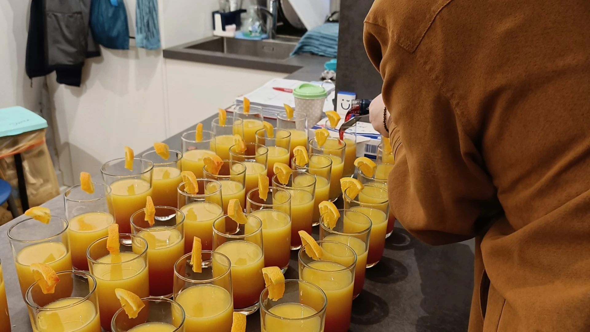 In einer K&uuml;che stehen Getr&auml;nkegl&auml;ser mit einem Cocktail und einer Orange dekoriert ordentlich angerichtet. Eine Frau ist mit dem Bef&uuml;llen der Gl&auml;ser besch&auml;ftigt.