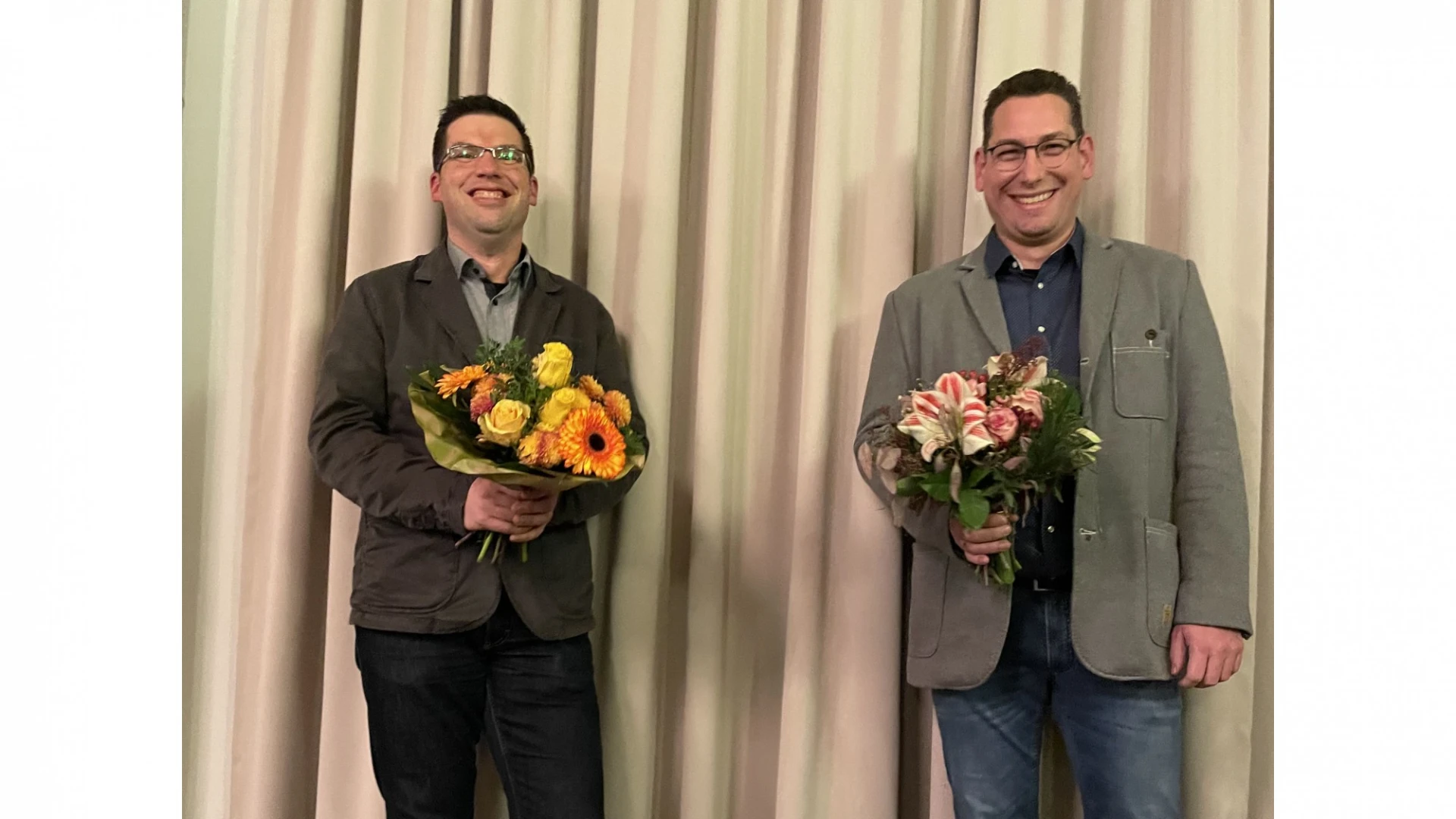 Zwei M&auml;nner stehen vor einem Vorhang mit einem Blumenstrau&szlig; in der Hand. Beide l&auml;cheln in die Kamera.