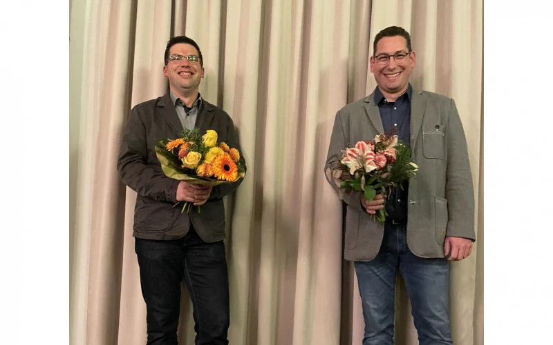 Zwei M&auml;nner stehen vor einem Vorhang mit einem Blumenstrau&szlig; in der Hand. Beide l&auml;cheln in die Kamera.