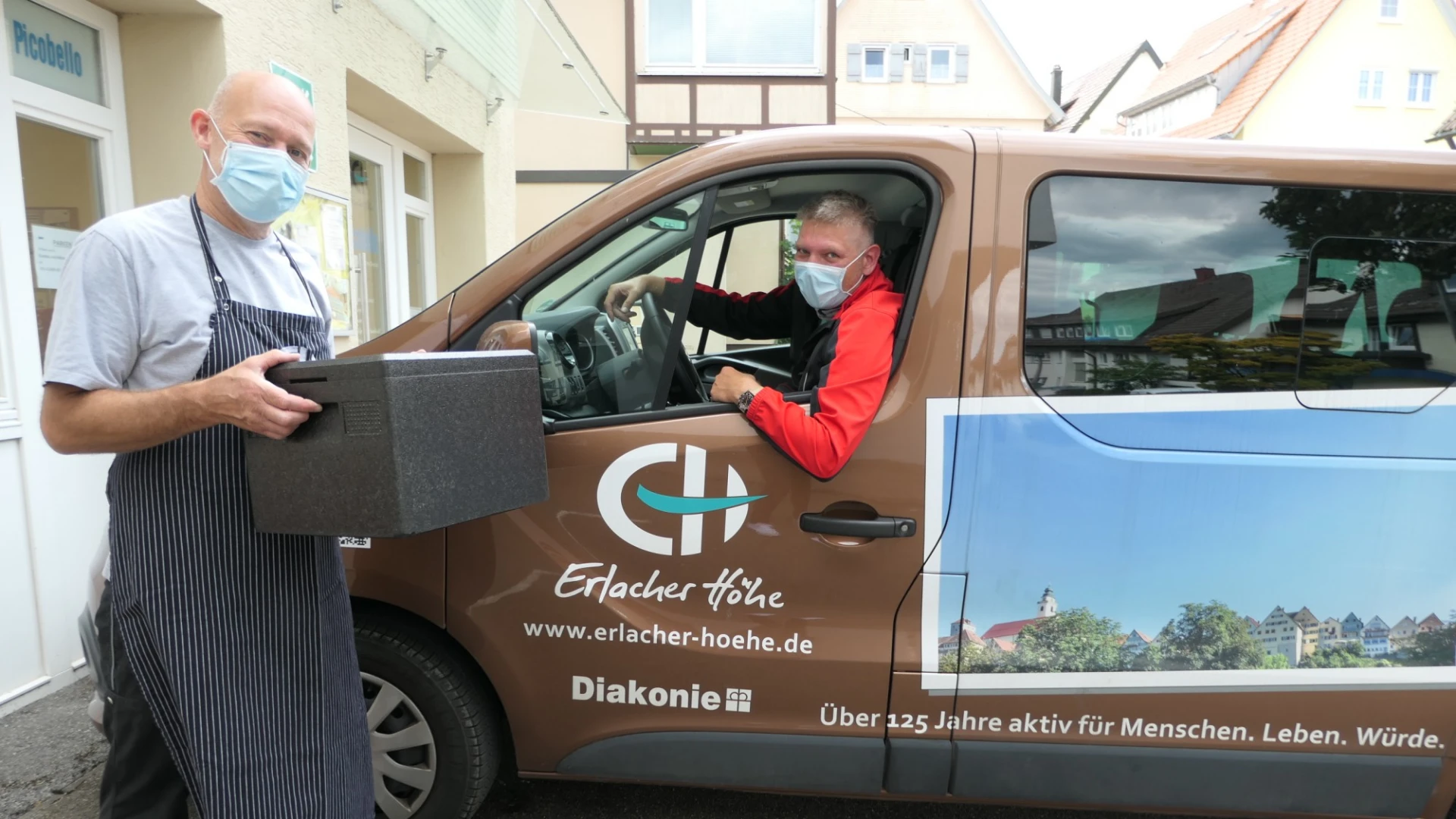 Ein Mann sitzt in einem braunen Lieferwagen, der mit dem Logo der Erlacher H&ouml;he beklebt ist. Neben ihm steht ein Mann mit Sch&uuml;rze und einer schwarzen Lieferbox. Beide tragen Mund-Nasenschutz.