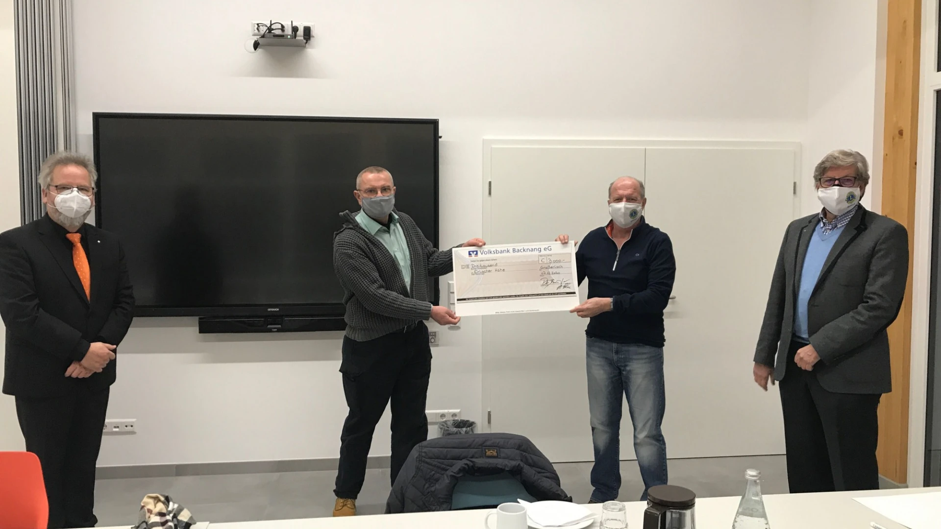 Vier M&auml;nner stehen in einem Raum vor einer wei&szlig;en Wand mit gro&szlig;em schwarzen Bildschirm. Alle tragen einen Mund-Nasenschutz. Die zwei M&auml;nner in der Mitte halten gemeinsam einen -Spendenscheck in der Hand. Vor ihnen befindet sich ein Konferenztisch mit Kaffeekanne, Wasserflasche, Gl&auml;ser, Geb&auml;ck und einer Brille.