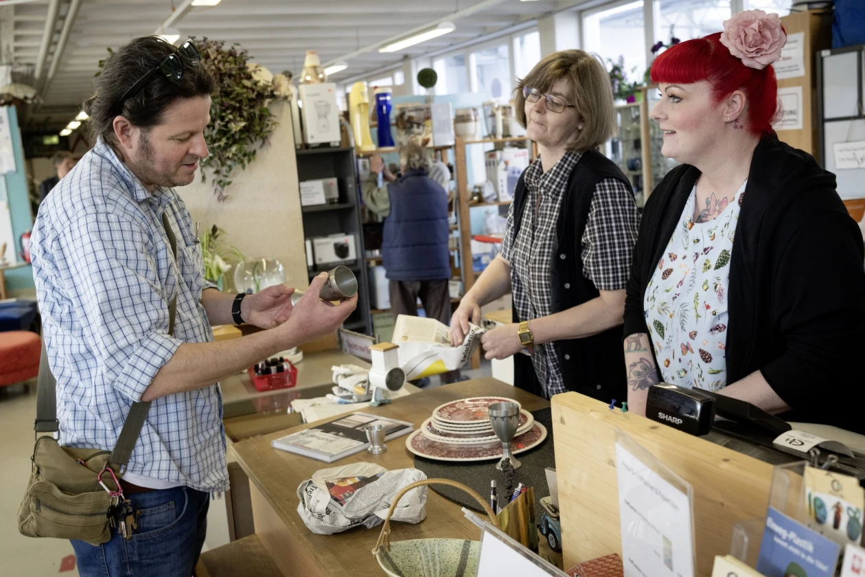 In einem Ladengesch&auml;ft h&auml;lt ein Mann einen Zinnbecher in der Hand und schaut ihn an. Hinter einer Verkaufstheke stehen zwei Frauen.