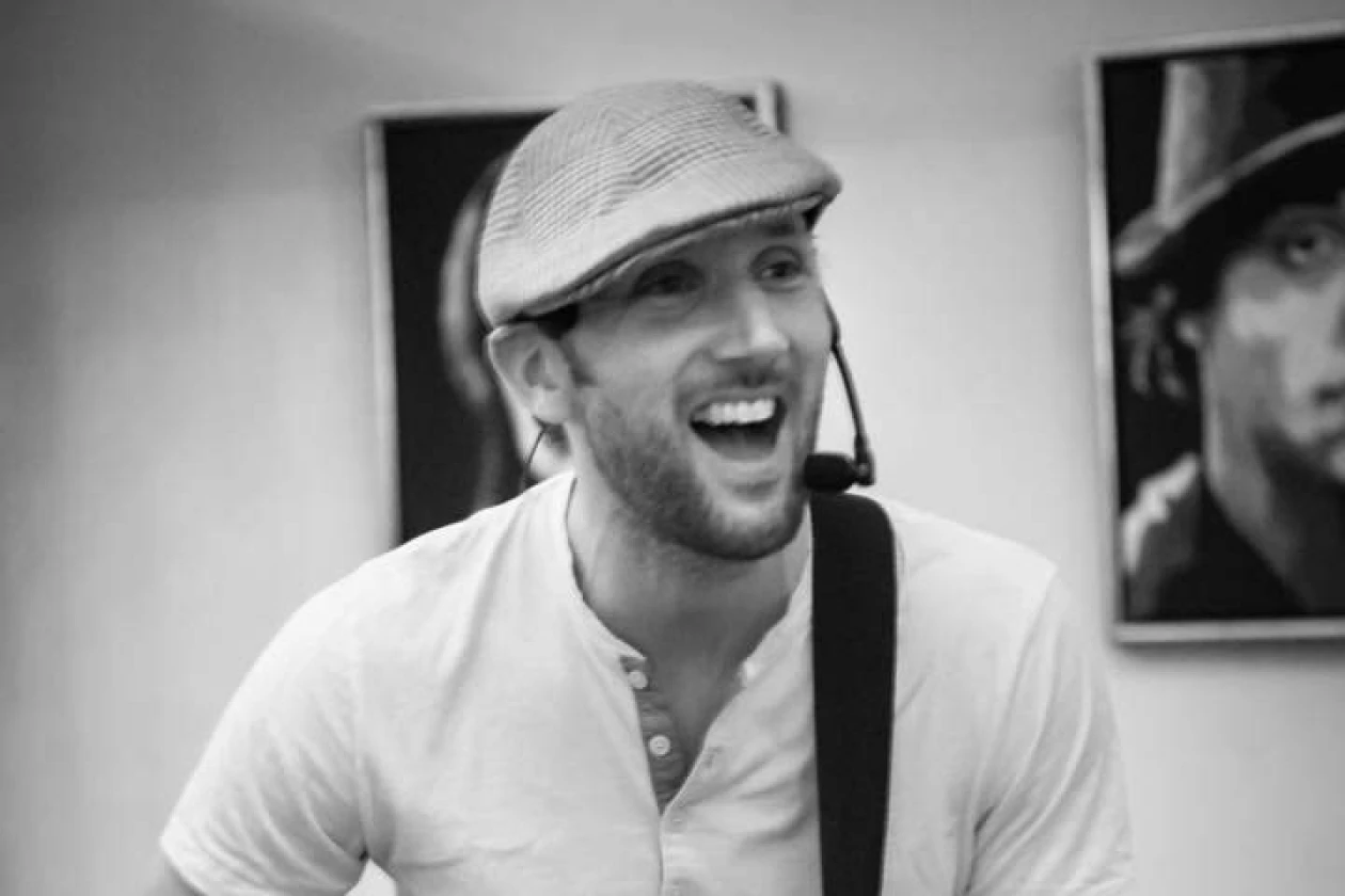 Auf einem Schwarz-Wei&szlig;-Bild ist ein Mann mit einer Gitarre zu sehen. Er tr&auml;gt eine M&uuml;tze und ein helles T-Shirt. Vor seinem Mund befindet sich ein Mikrofon. Im Hintergrund h&auml;ngen Bilder an einer hellen Wand.