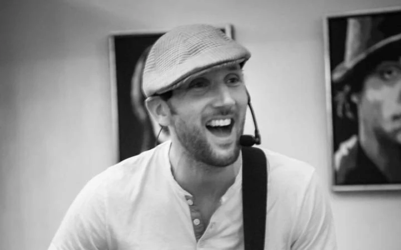 Auf einem Schwarz-Wei&szlig;-Bild ist ein Mann mit einer Gitarre zu sehen. Er tr&auml;gt eine M&uuml;tze und ein helles T-Shirt. Vor seinem Mund befindet sich ein Mikrofon. Im Hintergrund h&auml;ngen Bilder an einer hellen Wand.