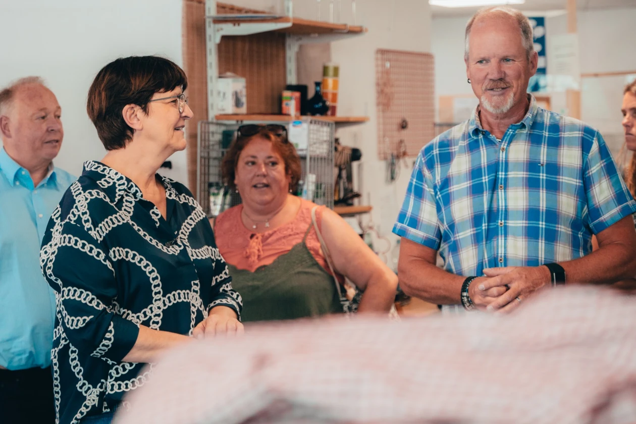 Mehrere Menschen mittleren Alters stehen in einem Raum, eine Frau in einem gemusterten Kleid spricht animiert, w&auml;hrend andere Personen zuh&ouml;ren. Im Vordergrund sind unscharfe Gegenst&auml;nde zu sehen, die Atmosph&auml;re wirkt lebendig und kommunikativ.