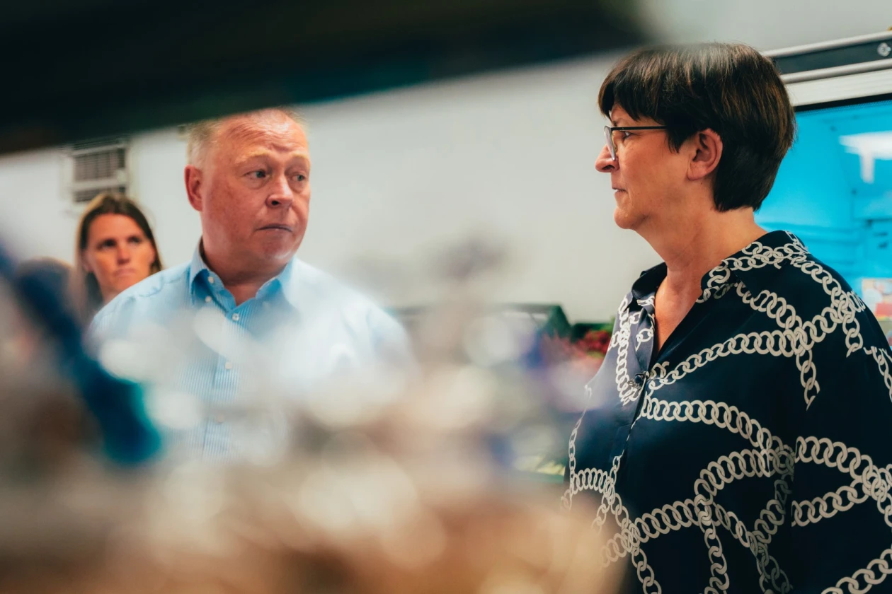 Eine Frau in einem gemusterten Kleid im Gespr&auml;ch mit einem Mann in hellblauem Hemd. Im Hintergrund ist eine weitere Frau zu sehen, sowie ein K&uuml;hlregal.