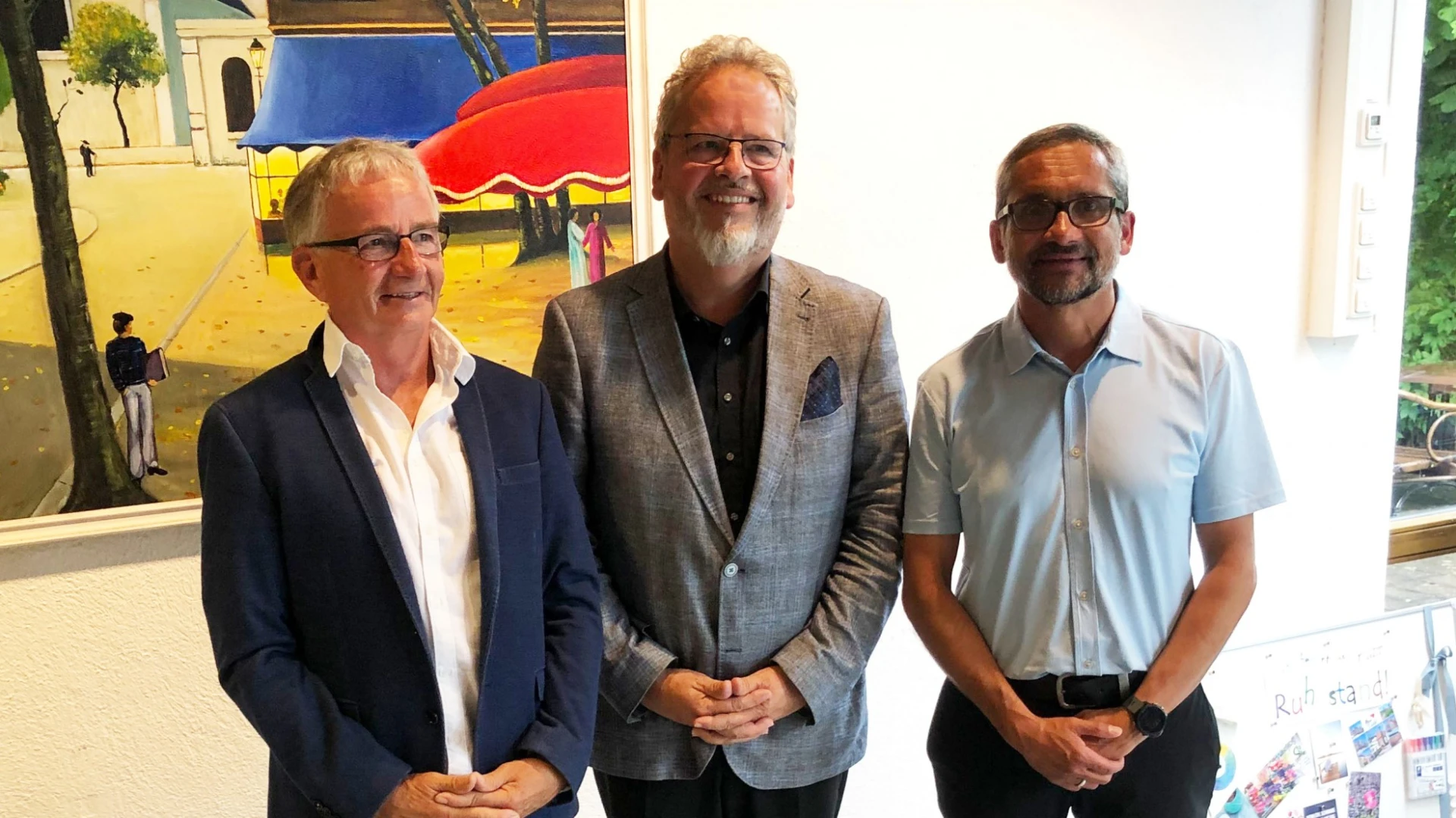 Drei M&auml;nner stehen nebeneinander. Ein &auml;lterer Mann mit wei&szlig;em Hemd und blauem Blazer, ein Mann mit lockigen grauen Haaren und Bart mit kariertem Jackett und ein j&uuml;ngerer Mann mit hellem Hemd und Brille. Im Hintergrund ist ein Gem&auml;lde zu sehen.