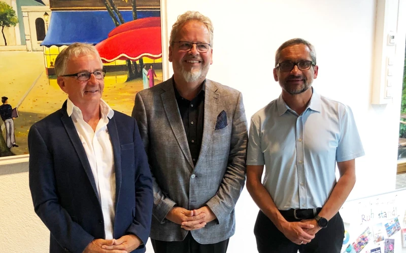 Drei M&auml;nner stehen nebeneinander. Ein &auml;lterer Mann mit wei&szlig;em Hemd und blauem Blazer, ein Mann mit lockigen grauen Haaren und Bart mit kariertem Jackett und ein j&uuml;ngerer Mann mit hellem Hemd und Brille. Im Hintergrund ist ein Gem&auml;lde zu sehen.