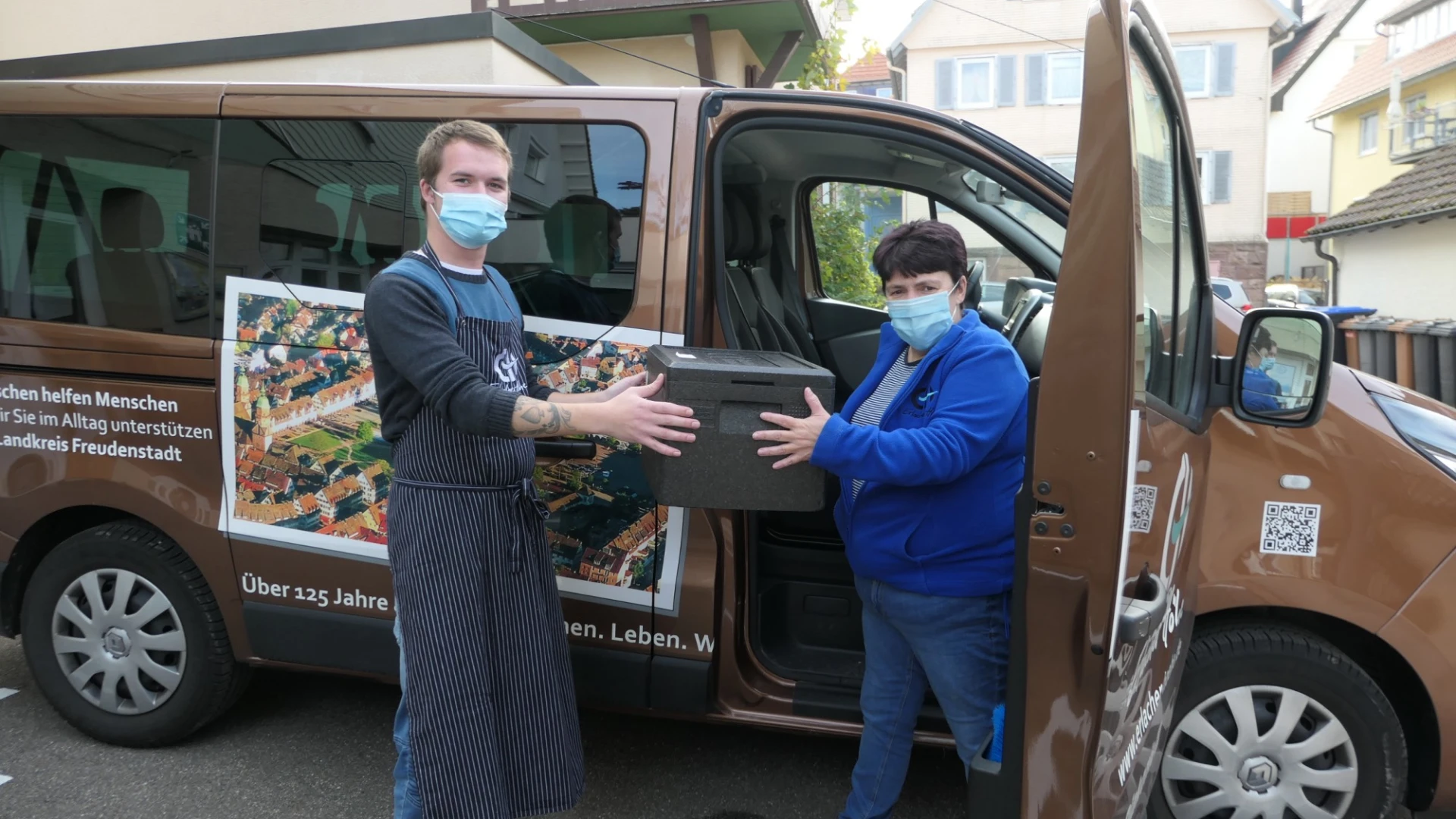 Ein Mann mit Sch&uuml;rze und eine Frau stehen vor einem braunen Transporter. Beide tragen medizinische Masken. Sie halten gemeinsam eine schwarze Kiste. Die Seitent&uuml;r des Fahrzeugs ist ge&ouml;ffnet.