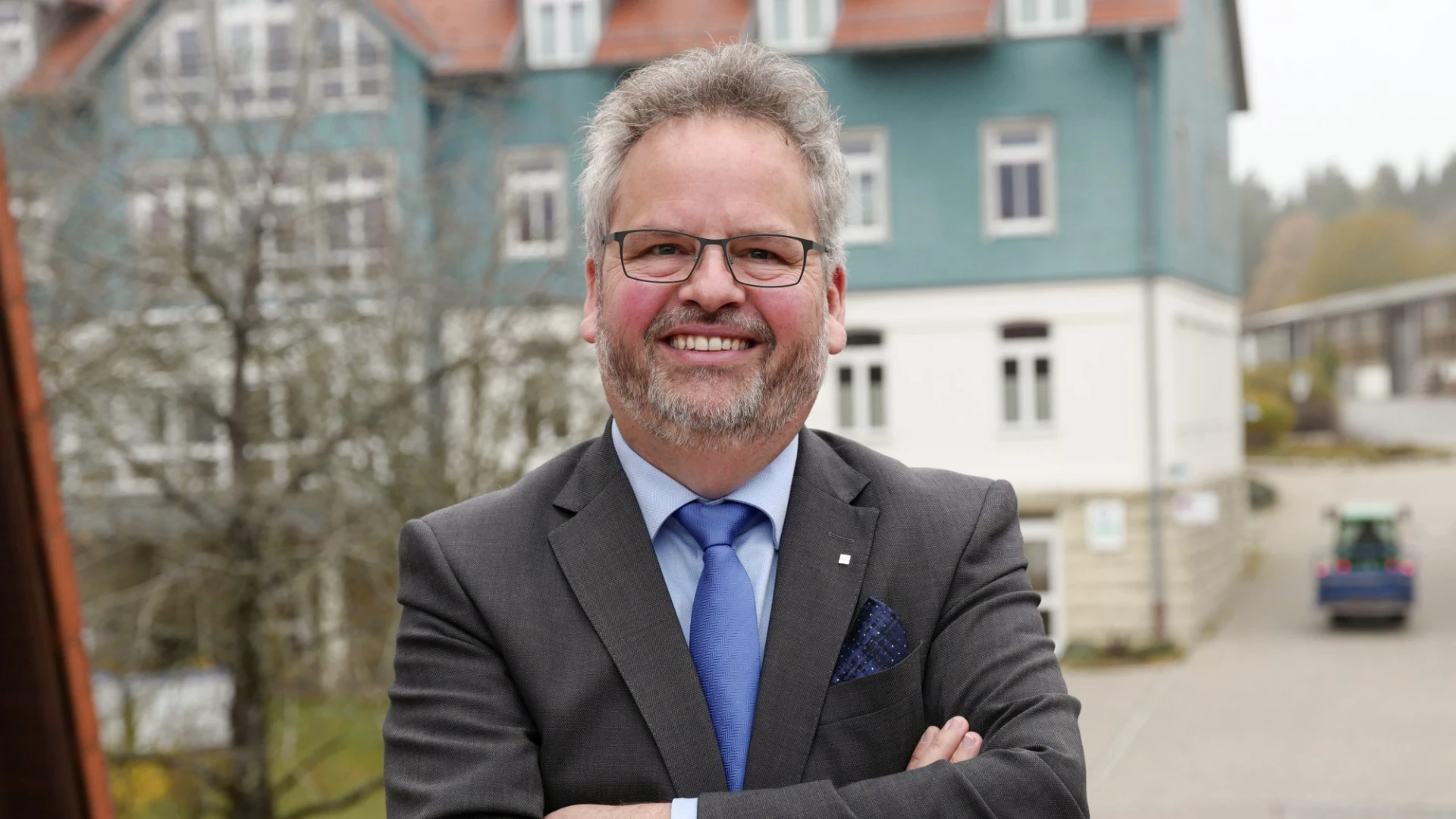 Ein &auml;lterer Mann mit grauen Haaren und Bart steht mit verchr&auml;nkten Armen selbstbewusst im Vordergrund eines Geb&auml;udes. Er tr&auml;gt einen grauen Anzug, eine blaue Krawatte und ein hellblaues Hemd, sowie eine Brille und l&auml;chelt in die Kamera.