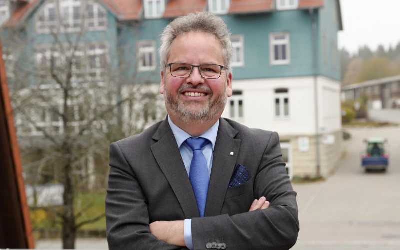 Ein &auml;lterer Mann mit grauen Haaren und Bart steht mit verchr&auml;nkten Armen selbstbewusst im Vordergrund eines Geb&auml;udes. Er tr&auml;gt einen grauen Anzug, eine blaue Krawatte und ein hellblaues Hemd, sowie eine Brille und l&auml;chelt in die Kamera.