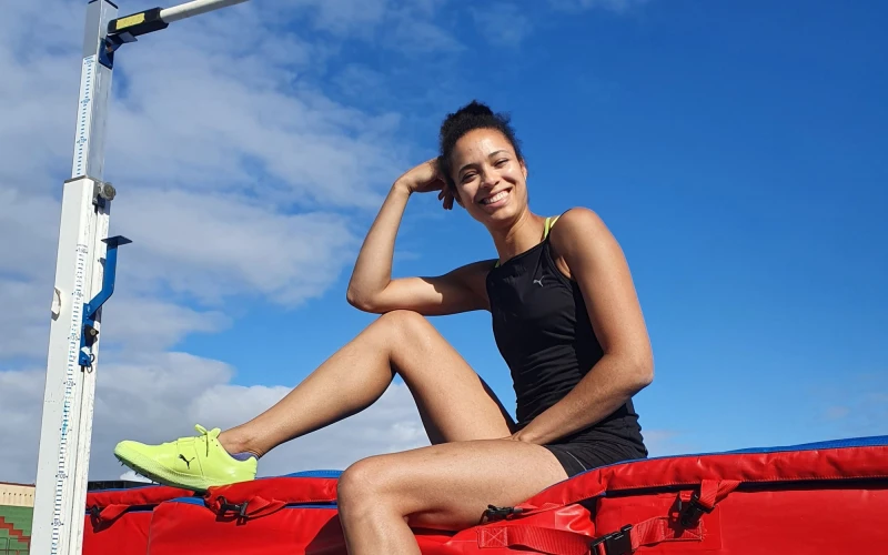 Eine junge Hochspringerin in dunkler Sportkleidung mit gelben Sportschuhen sitzt auf einer roten Matte unter der Hochsprunglatte und l&auml;chelt in die Kamera.