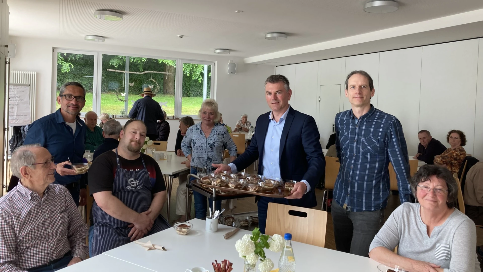 Ein Mann mittleren Alters in hellblauem Hemd und dunklem Jackett serviert beim Mittagstisch Dessert in Sch&auml;lchen. Ein weitere Mann steht links und ein weiterer Mann rechts von ihm, beide  freuen sich &uuml;ber die Unterst&uuml;tzung.