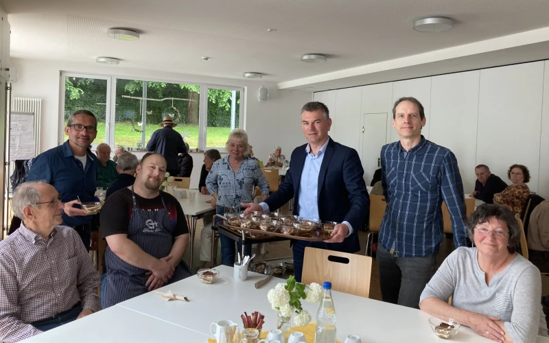 Ein Mann mittleren Alters in hellblauem Hemd und dunklem Jackett serviert beim Mittagstisch Dessert in Sch&auml;lchen. Ein weitere Mann steht links und ein weiterer Mann rechts von ihm, beide  freuen sich &uuml;ber die Unterst&uuml;tzung.