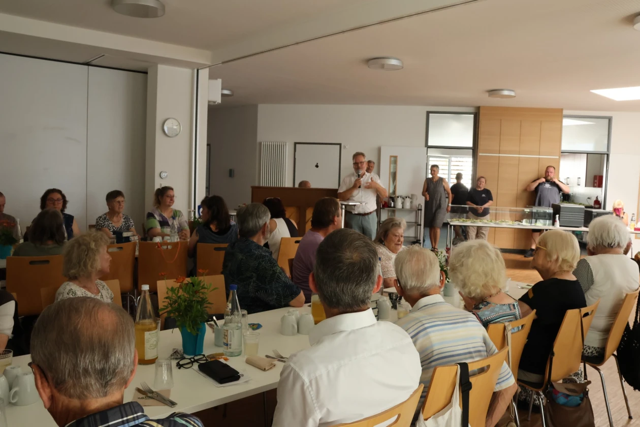 Eine Gruppe von Menschen sitzt in einem hellen Seminarraum auf Holzst&uuml;hlen an Tischen, w&auml;hrend eine Person vorne steht und zu den Besuchern spricht