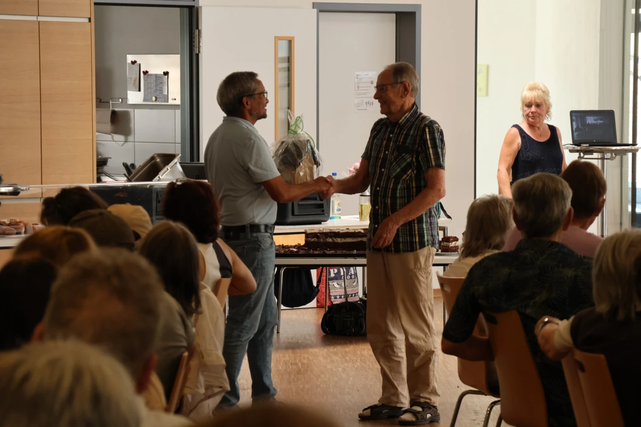 Zwei M&auml;nner sch&uuml;tteln sich die Hand vor einer Gruppe von Besuchern in einem Schulungsraum