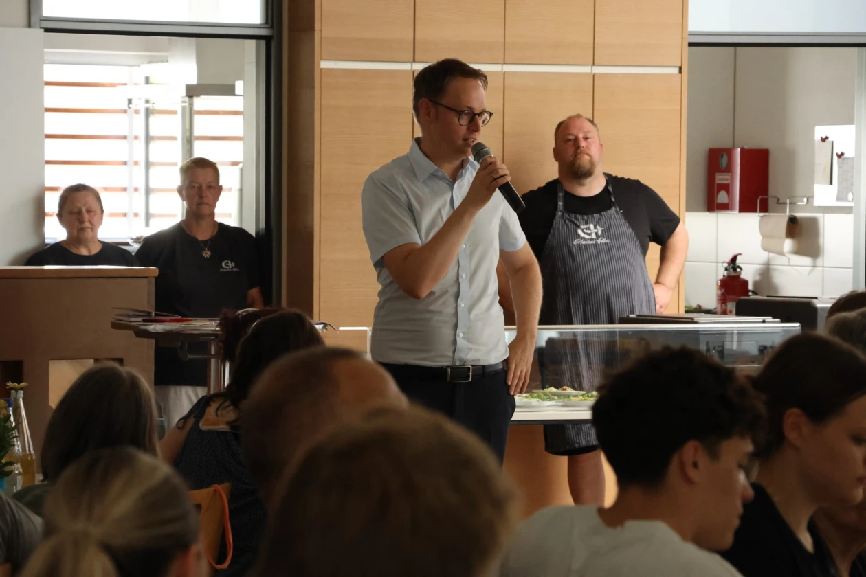 Ein Mann mit Brille und Mikrofon spricht vor einer Gruppe von Zuh&ouml;rern in einem hellen Raum