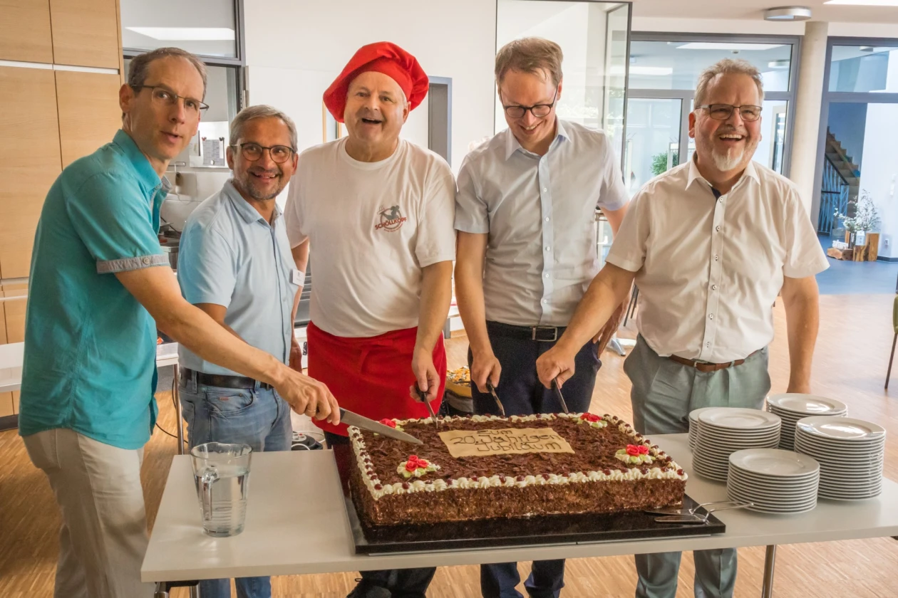 F&uuml;nf M&auml;nner stehen l&auml;chelnd hinter einem gro&szlig;en Schokoladenkuchen auf einem Tisch