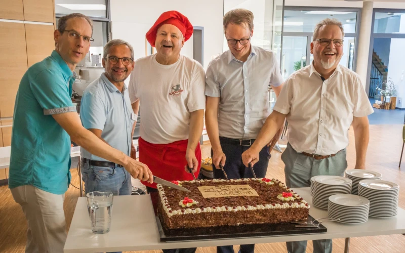 F&uuml;nf M&auml;nner stehen l&auml;chelnd hinter einem gro&szlig;en Schokoladenkuchen auf einem Tisch