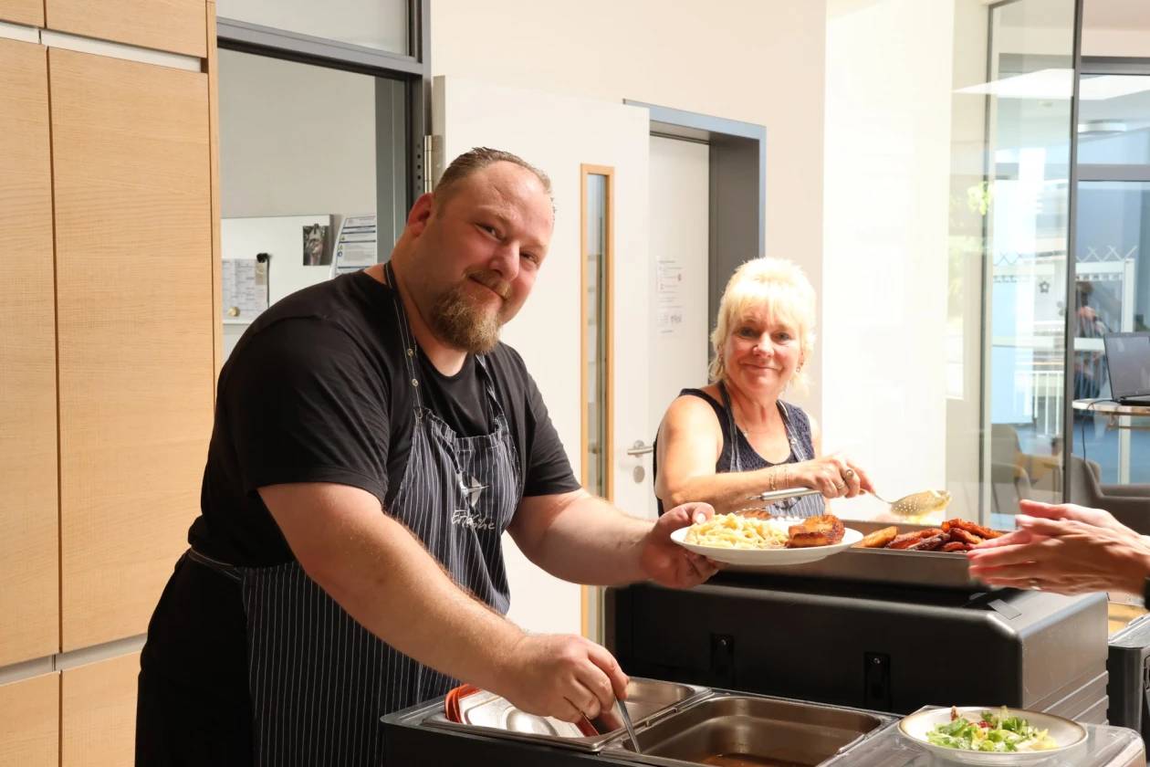 Ein Mann in schwarzem T-Shirt und Sch&uuml;rze serviert Speisen an einem Buffet, w&auml;hrend eine Frau mit blonden Haaren neben ihm steht