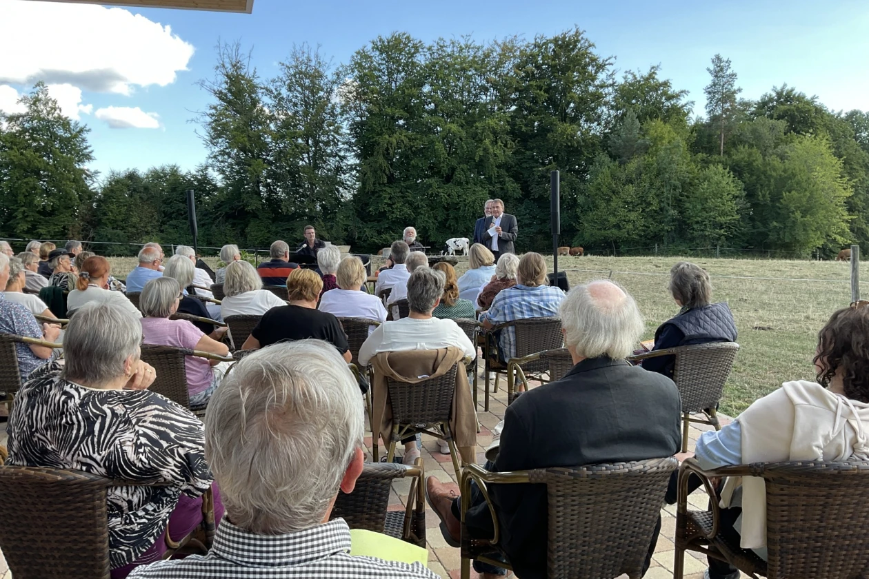 Menschen sitzen in Gartenst&uuml;hlen auf einer Terrasse und blicken zu einem Mann, der zu ihnen redet. Im Hintergrund B&auml;ume und blauer Himmel mit wei&szlig;en Wolken.
