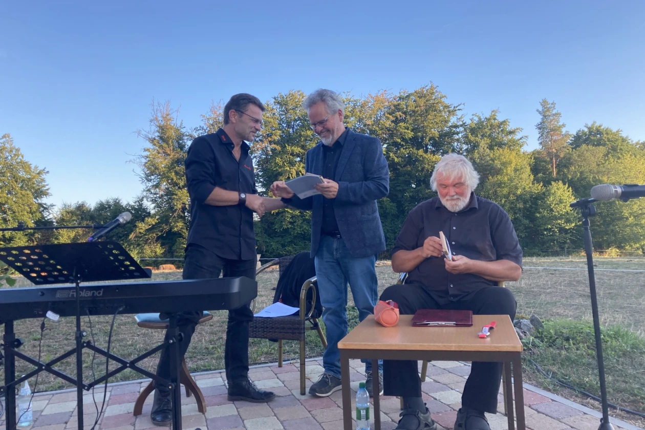 Drei M&auml;nner auf einer Terrasse mit B&auml;umen im Hintergrund. Ein &auml;lterer Mann sitzt am Tisch, w&auml;hrend zwei andere M&auml;nner im Gespr&auml;ch stehen. Die Szene wirkt ruhig und entspannt an einem sonnigen Tag.