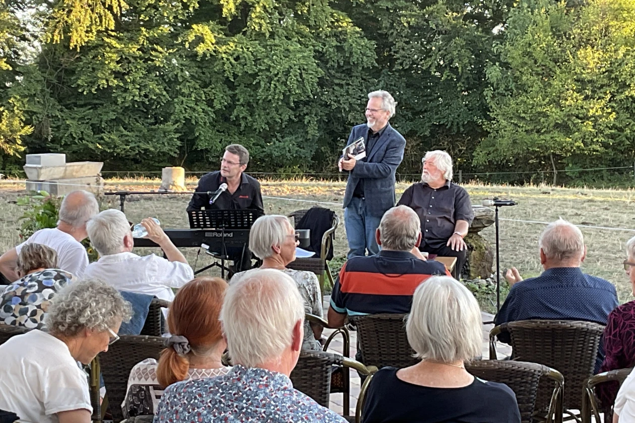 Menschen auf Gartenst&uuml;hlen bei einer Veranstaltung im Freien h&ouml;ren einem Redner zu. Im Hintergrund sind zwei weitere M&auml;nner zu sehen, einer mit Mirkofon, einer am E-Piano.