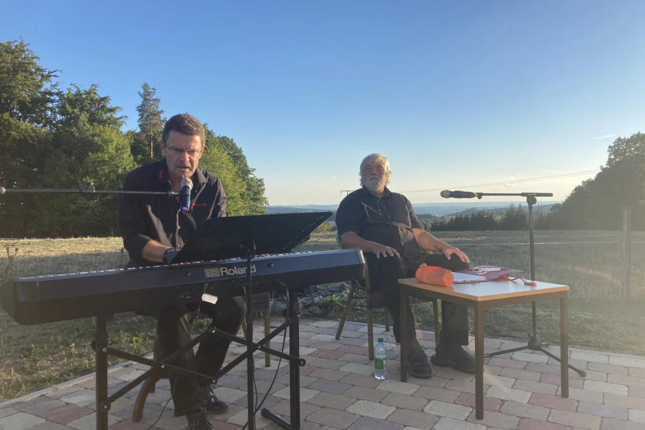 Ein Mann in dunkler Kleidung sitzt an einem E-Piano, ein weiterer Mann in dunkler Kleidung im Hintergrund an einem kleinen Tisch mit Mikrofon. Die Veranstaltung findet im Freien statt. Im Hintergrund ist Wiesenfl&auml;che und Wald, sowie der Horizont zu sehen.