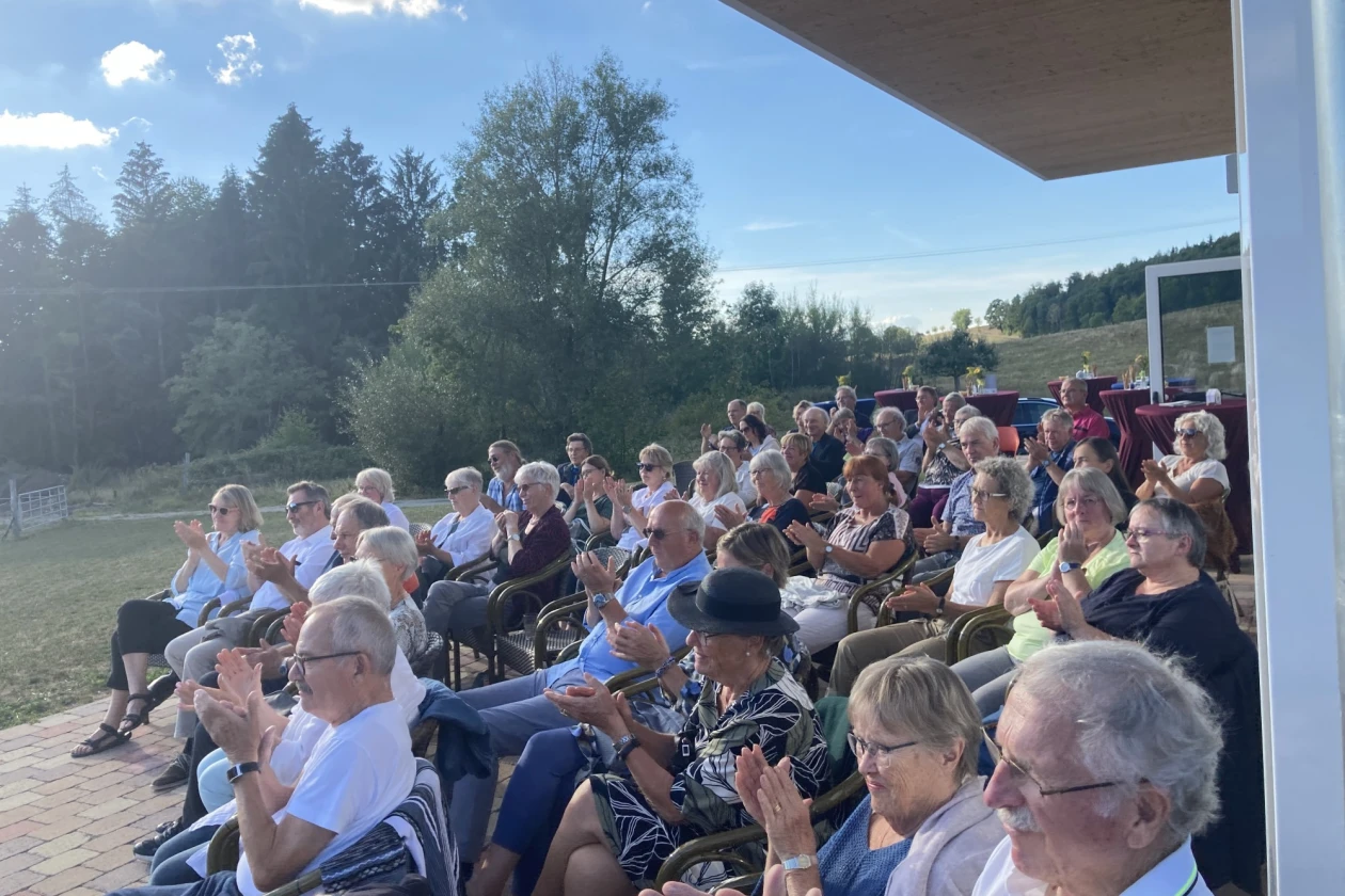 Publikum bei einer Veranstaltung im Freien, das auf St&uuml;hlen sitzt und eine Pr&auml;sentation oder Veranstaltung verfolgt, vor einer Kulisse von B&auml;umen und blauem Himmel.