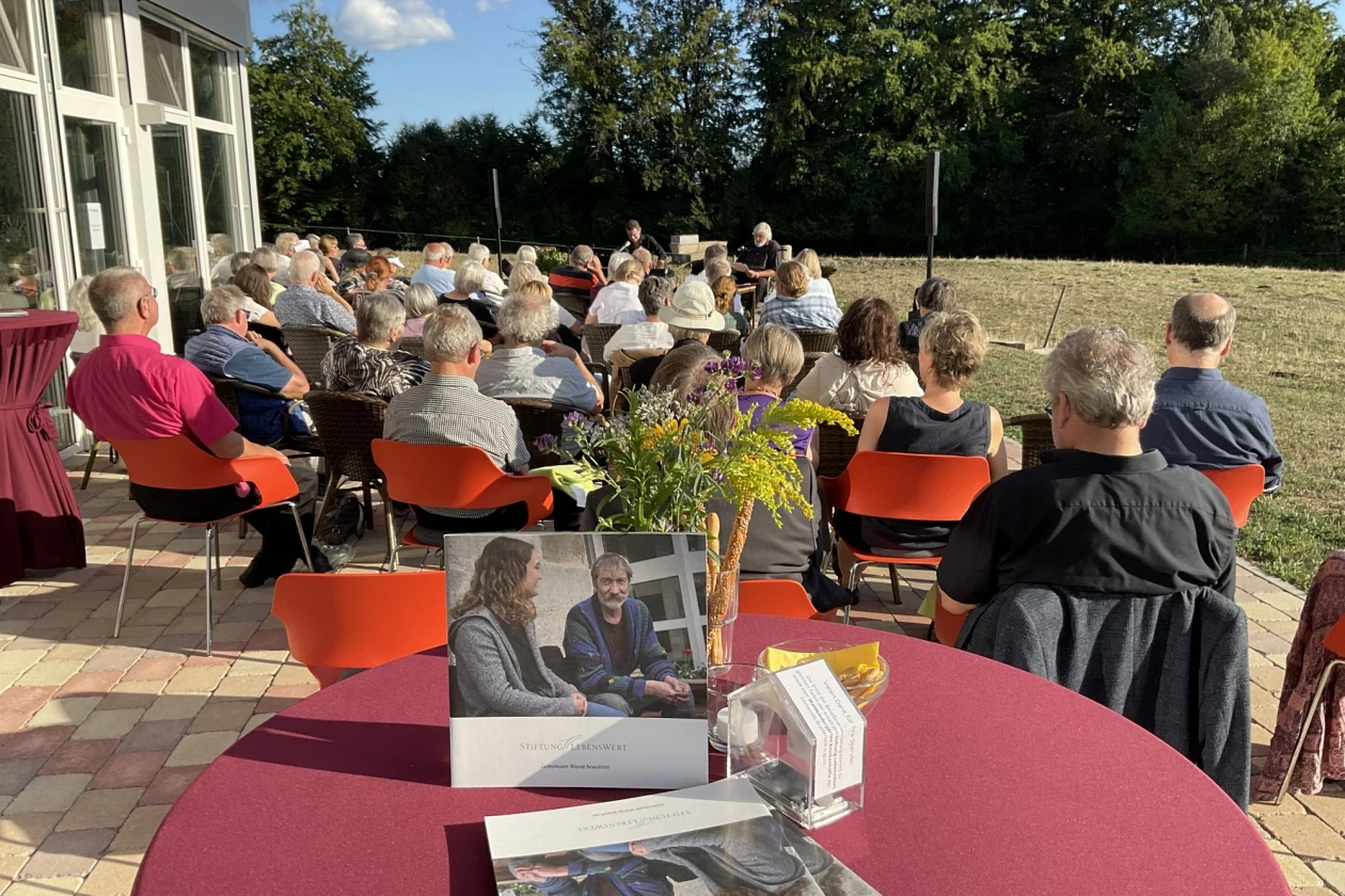 Mehrere Menschen sitzen auf einer Terrasse mit roten und orangefarbenen St&uuml;hlen, umgeben von B&auml;umen und Gr&uuml;nfl&auml;chen, und nehmen an einer Veranstaltung oder Besprechung im Freien teil.