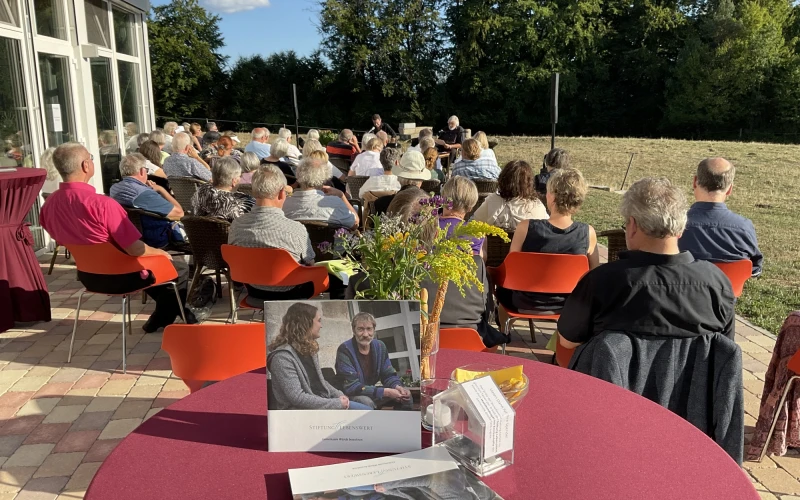 Mehrere Menschen sitzen auf einer Terrasse mit roten und orangefarbenen St&uuml;hlen, umgeben von B&auml;umen und Gr&uuml;nfl&auml;chen, und nehmen an einer Veranstaltung oder Besprechung im Freien teil.