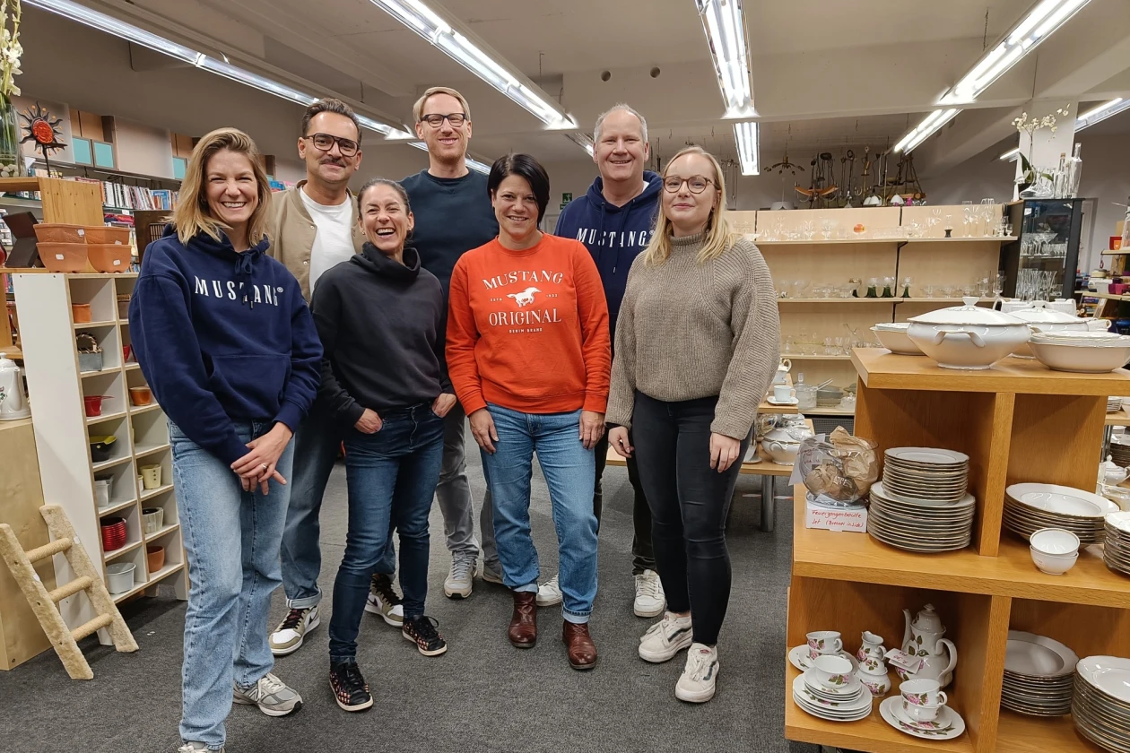 Sieben Personen stehen in einem Raum, in dem sich links und rechts Regale mit Geschirr, Tassen und Porzellan befinden. Im Hintergrund sind weitere Regale zu sehen.