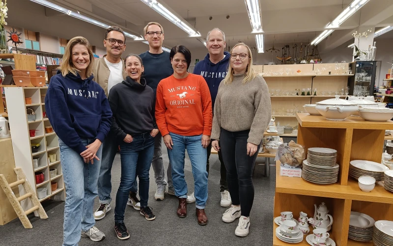 Sieben Personen stehen in einem Raum, in dem sich links und rechts Regale mit Geschirr, Tassen und Porzellan befinden. Im Hintergrund sind weitere Regale zu sehen.