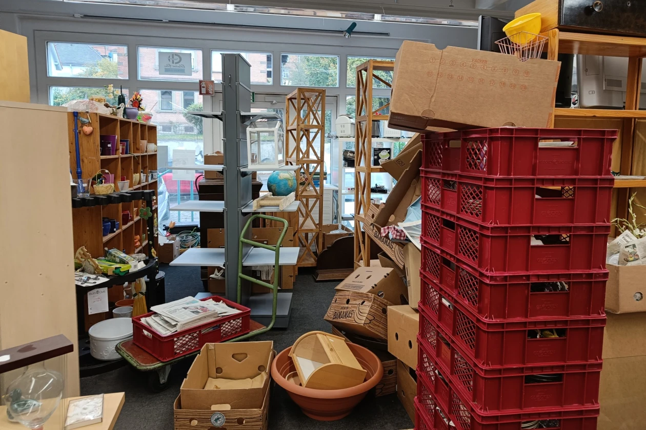 Ein Raum mit zahlreichen Regalen, Holzm&ouml;beln und Stapeln roter Kunststoffkisten ist zu sehen. In der Mitte liegen Kartons und K&ouml;rbe verstreut.