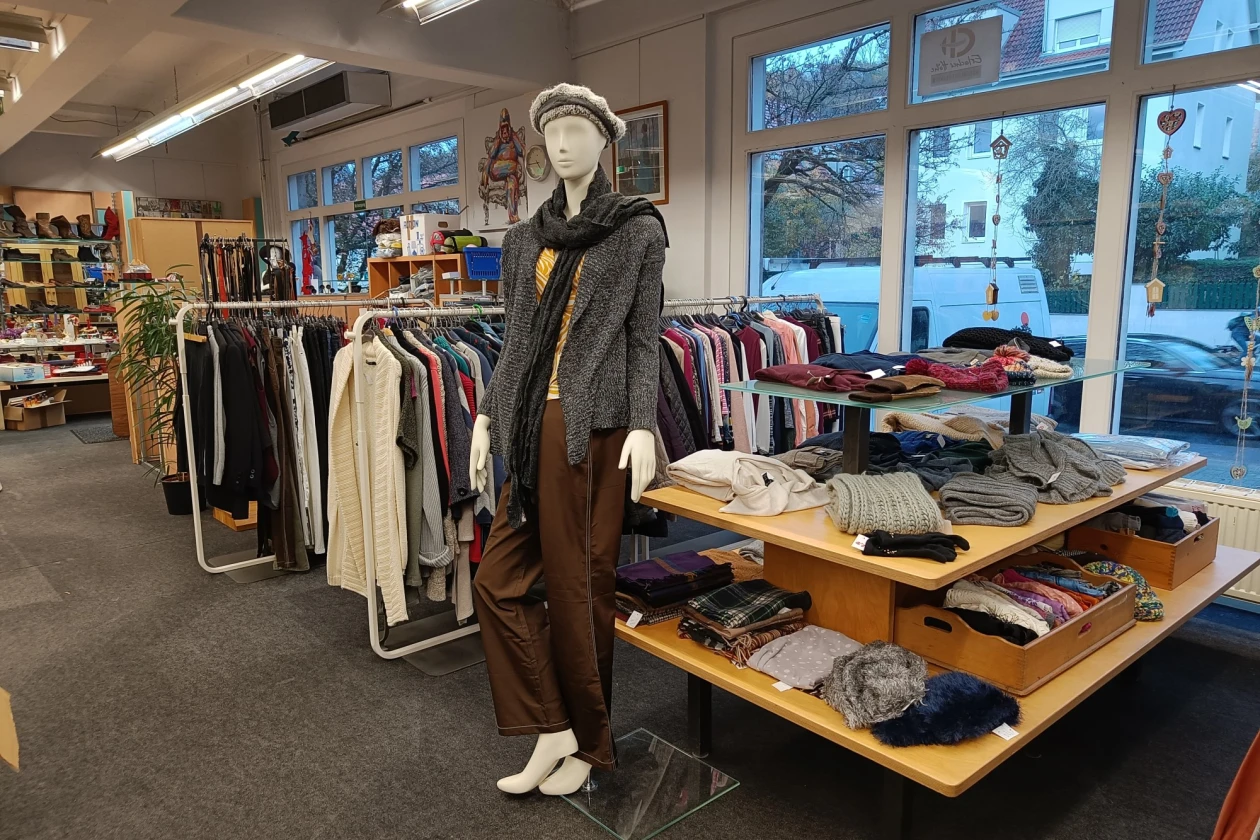 Zu sehen ist ein Verkaufsraum. Im Vordergrund steht eine Schaufensterpuppe, gekleidet in einer braunen Hose, einem gelben Oberteil, einer grauen Jacke und einem Schal. Im Hintergrund befinden sich Kleiderst&auml;nder und Auslagen mit verschiedenen Kleidungsst&uuml;cken.