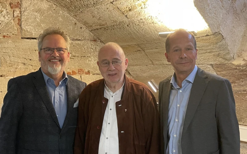 Drei M&auml;nner stehen mit Hemd und Jacketts in einem Kellergew&ouml;lbe. Sie blicken l&auml;chelnd in die Kamera.