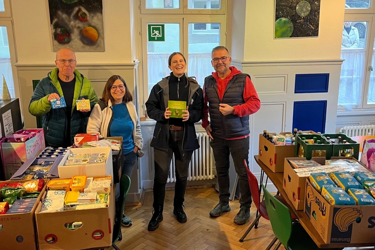 Vier Personen stehen in einem Raum mit Spendendosen und Kartons voller Lebensmittel und Waren