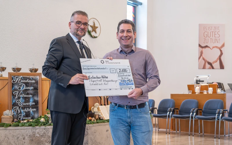 Zwei M&auml;nner halten gemeinsam einen Spendenscheck &uuml;ber 2.600 Euro in einem Gemeinderaum