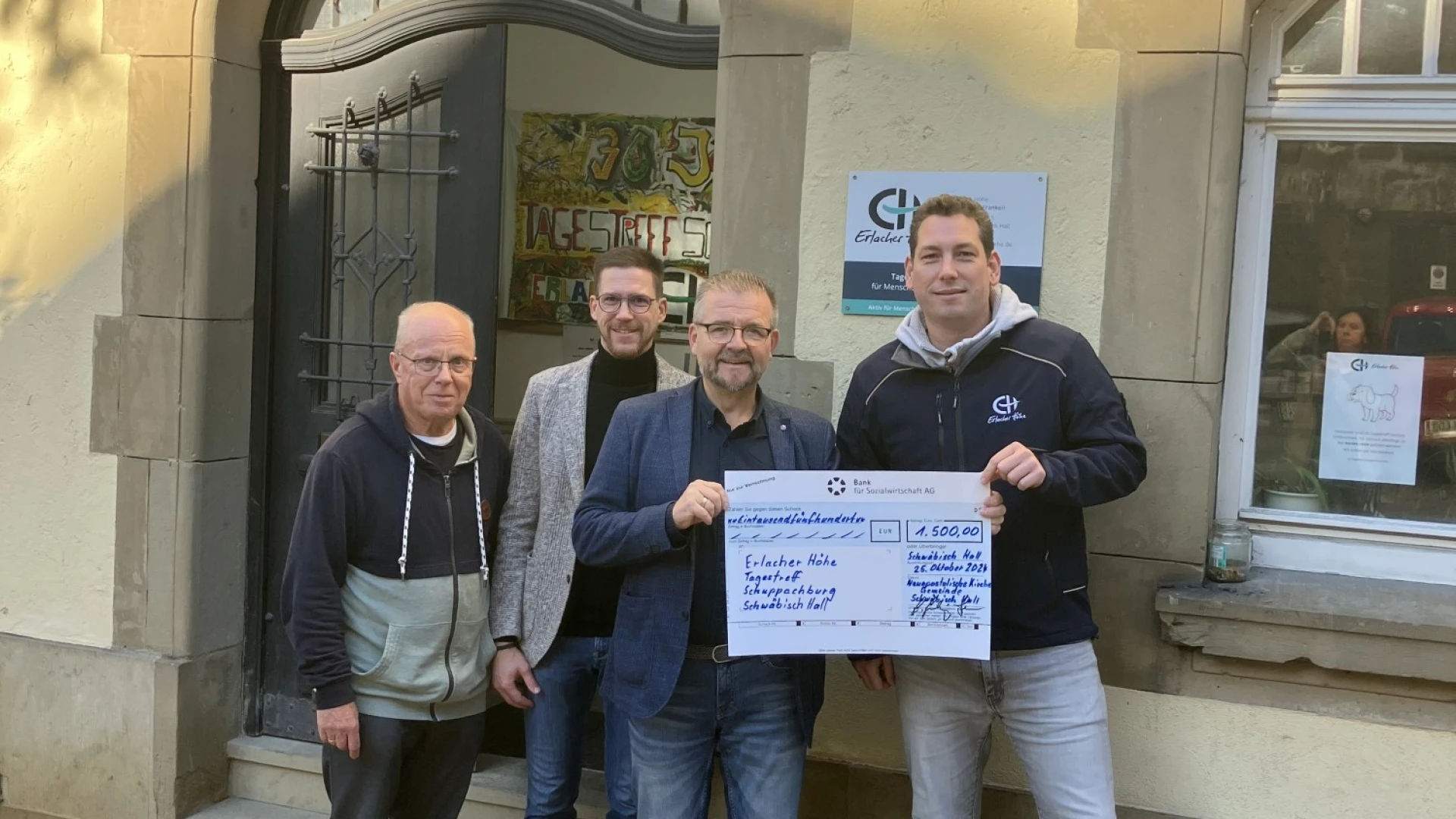 Vier M&auml;nner stehen vor einem Geb&auml;ude bei einer Scheck&uuml;bergabe. Alle l&auml;cheln freundlich in die Kamera.