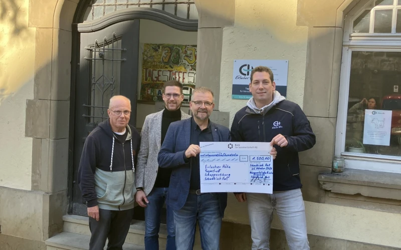 Vier M&auml;nner stehen vor einem Geb&auml;ude bei einer Scheck&uuml;bergabe. Alle l&auml;cheln freundlich in die Kamera.