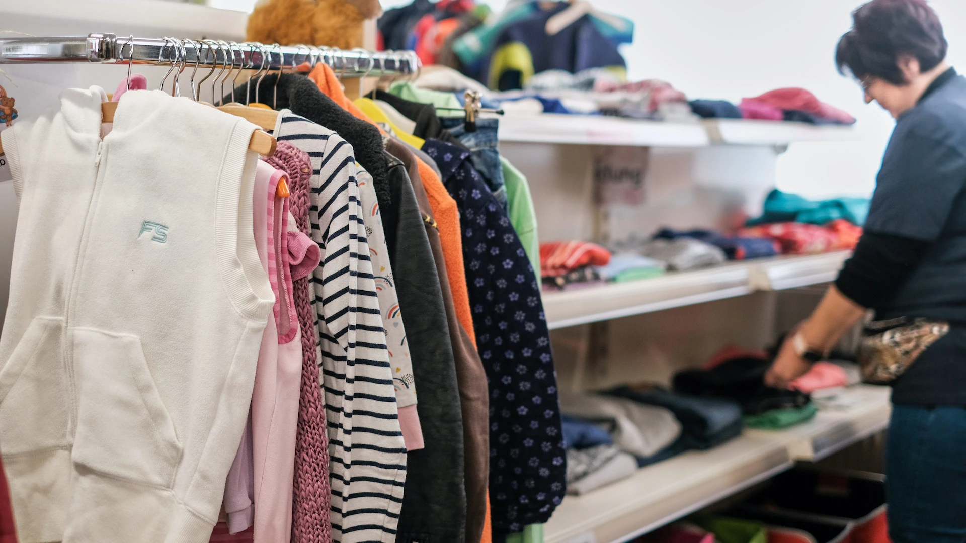 Kleiderst&auml;nder mit verschiedenfarbigen Kleidungsst&uuml;cken in einem Ladengesch&auml;ft. Eine Frau steht im Hintergrund und sortiert Textilien in Regalen.