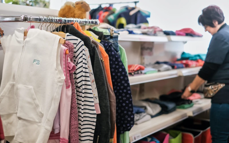 Kleiderst&auml;nder mit verschiedenfarbigen Kleidungsst&uuml;cken in einem Ladengesch&auml;ft. Eine Frau steht im Hintergrund und sortiert Textilien in Regalen.