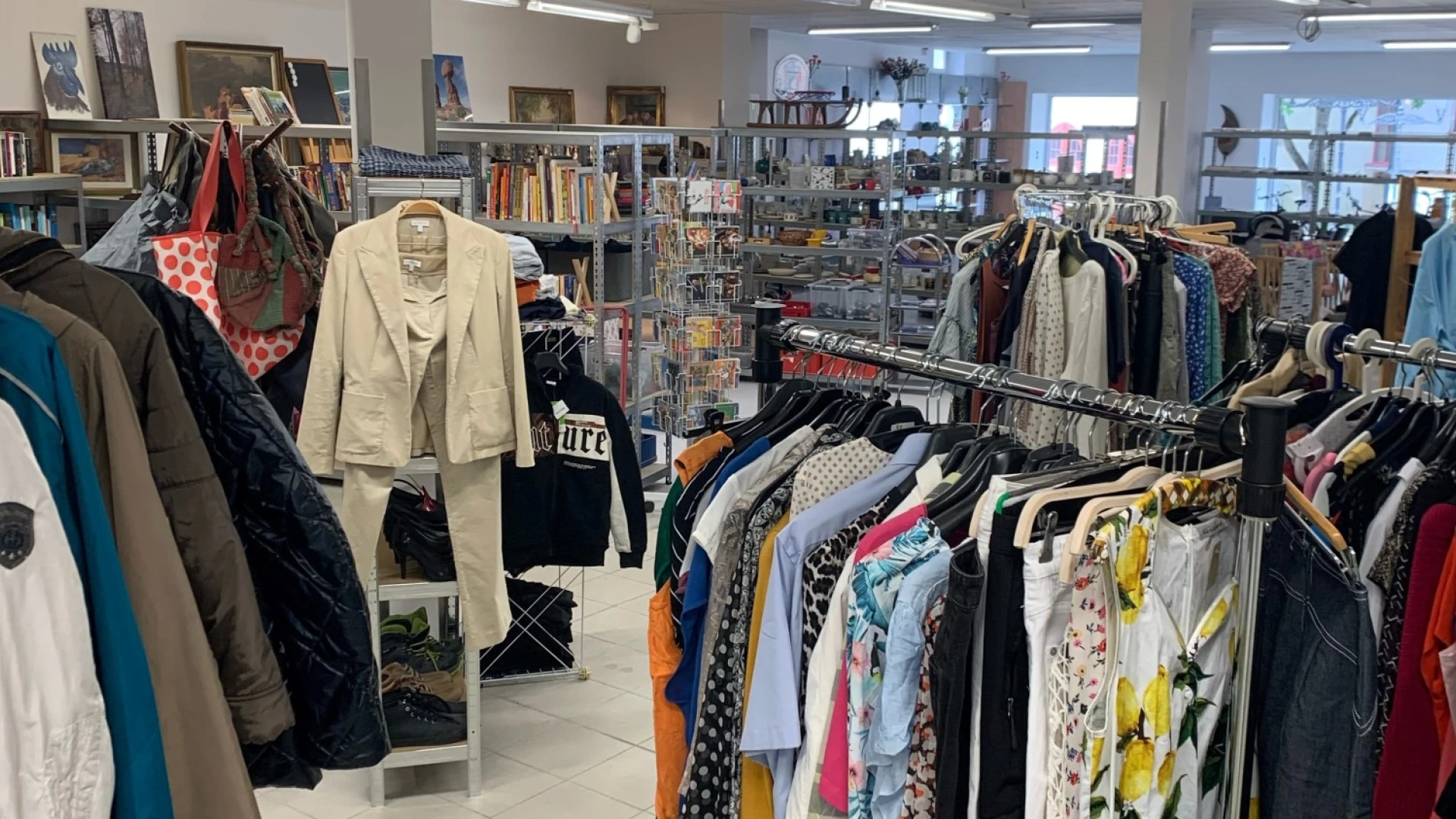 Helles, ger&auml;umiges Ladengesch&auml;ft mit Kleiderst&auml;ndern voller bunter Jacken, Hemden und Kleider. Regale mit B&uuml;chern und Accessoires stehen im Hintergrund. Wei&szlig;e Bodenfliesen und Deckenbeleuchtung pr&auml;gen den freundlichen Raum.