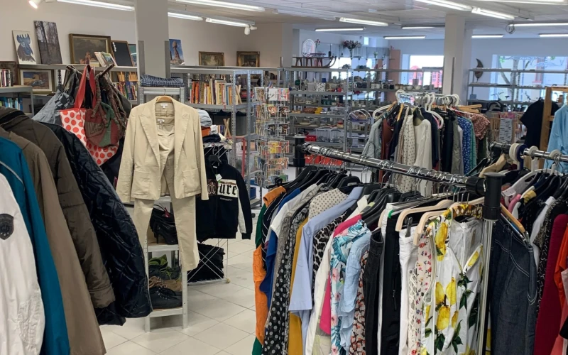 Helles, ger&auml;umiges Ladengesch&auml;ft mit Kleiderst&auml;ndern voller bunter Jacken, Hemden und Kleider. Regale mit B&uuml;chern und Accessoires stehen im Hintergrund. Wei&szlig;e Bodenfliesen und Deckenbeleuchtung pr&auml;gen den freundlichen Raum.